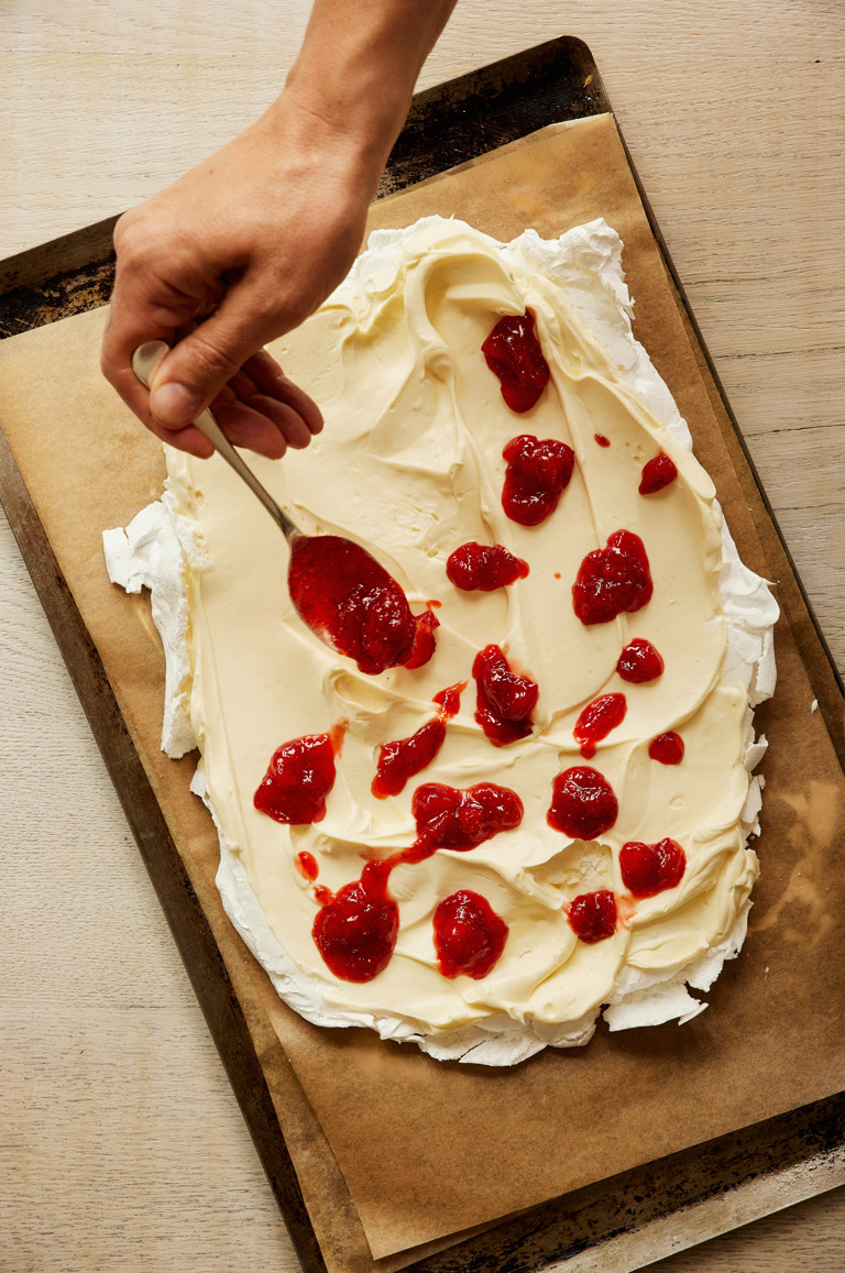 A close-up portrait shot of the process of making the strawberry and lime meringue roulade: the meringue is being topped with cream and strawberry compote