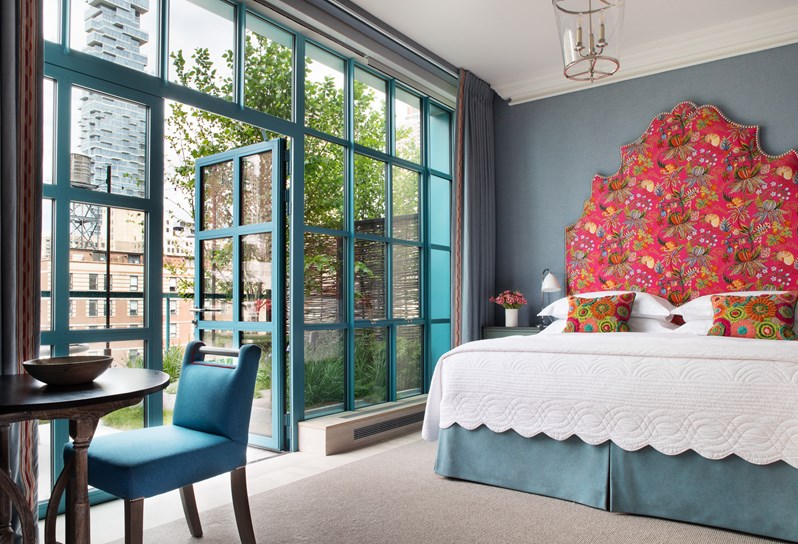 A bedroom with floor to ceiling doors that open up onto a landscaped terrace. There is a bed with a pink patterned headboard and a bedside table. In front there is a small wooden table with a blue chair.