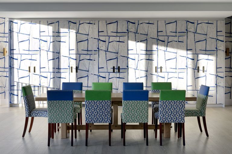 A view of the entertaining area on the top floor featuring a large wooden dining table and 10 chairs upholstered in colourful patterned fabrics. Behind the table are floor-to-ceiling cupboards covered in a graphic, geometric print at 9-11 Richmond Buildings