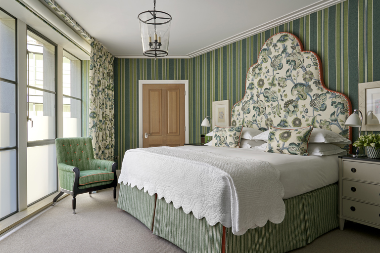 A diagonal shot of the bedroom in the Deluxe Two Bedroom Suite room 5 at Ham Yard Hotel. With a stylish bedroom featuring a vibrant patterned headboard and pillows, green textured wallpaper, eclectic artwork and floral arrangement.