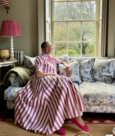 A press image of Paula Sutton. Paula is sat on a blue sofa looking at the camera in a pink and white striped dress.