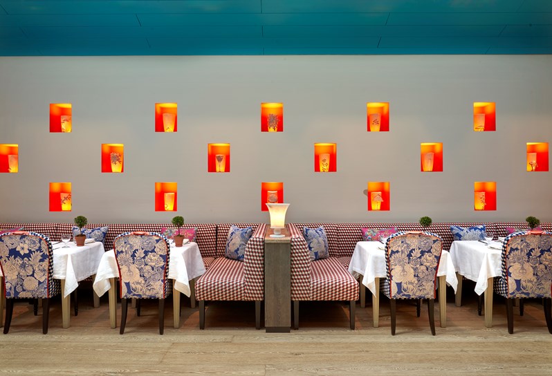 A wall showcasing a light installation of illuminated ceramic pots. In front there is a stretch of red and white patterned banquette seating, with tables of two set up in front. On the other side of each table is a blue and white floral dining chair.