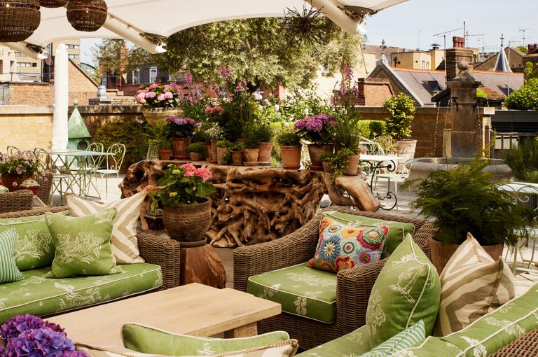 A wide angle shot of the roof terrace at Ham Yard Hotel, featuring sofa seating, dining tables, floral arrangements and the fountain