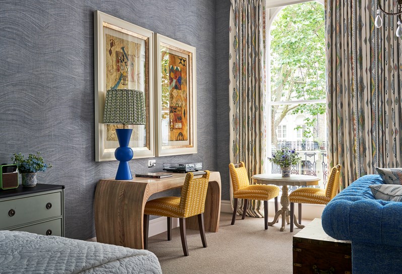 Seating area shot of room 101, the Knightsbridge Suite at Knightsbridge Hotel. image shows a diagonal shot from the back of the sofa area looking towards the far left window with dining area of small circular table with two yellow chairs next to a large desk with a yellow chair and blue statement lamp on top.