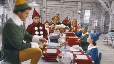 A still from 'Elf'. A long work table with elves dressed in different coloured outfits sitting to make toys. At the end, a large elf can be seen sat on a small stool as the others look at him