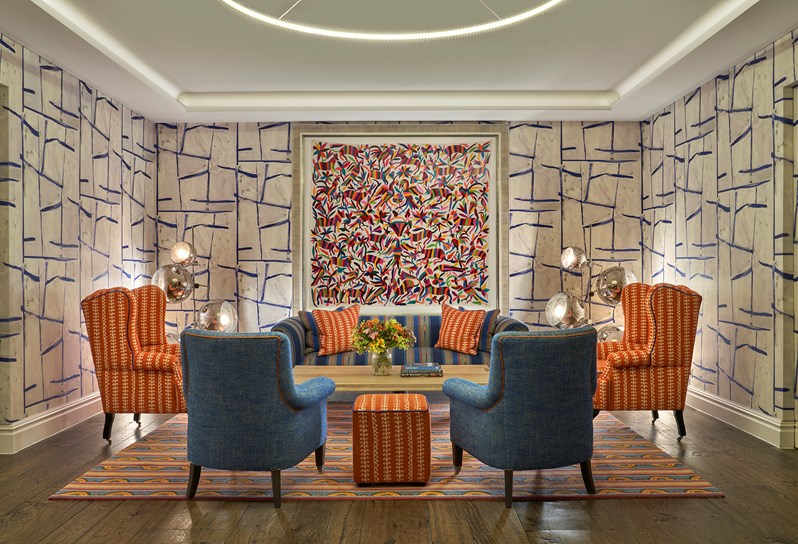 An image of the seating area in the Sandra Blow private events room at The Soho Hotel. The image shows a sofa in a blue and orange colour scheme under a piece of art, with two orange armchairs flanking the sofa and two blue arm chairs at the front facing away from the camera.