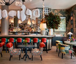 The bar at Warren Stree Hotel in Tribeca. Red and teal leather bar stools stand at a pewter-topped bar. Overhead hangs an abstract sculpture in white and gold that runs the length of the bar.