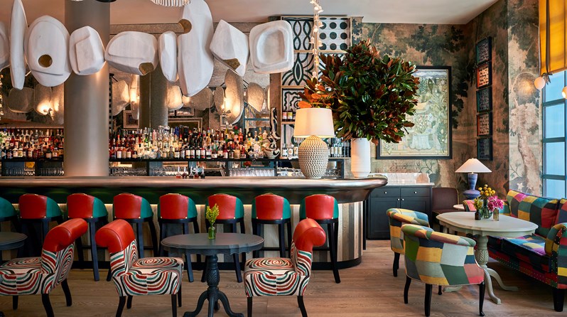 The bar at Warren Stree Hotel in Tribeca. Red and teal leather bar stools stand at a pewter-topped bar. Overhead hangs an abstract sculpture in white and gold that runs the length of the bar.