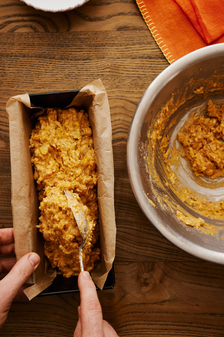 In this image you can see hand filling a loaf tin with the cornbread mixture. on the right side of the image we see the bowl in which the mixture was made almost empty. 