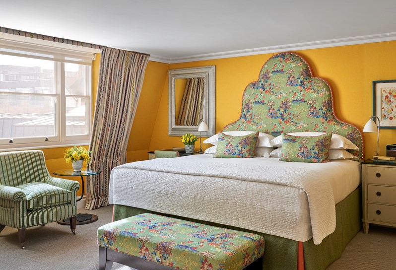 A yellow bedroom with a floral headboard. There are 2 lamps either side of the bed on the bedside tables. A desk and chair with a mirror hanging above. There is a ottoman at the end of the bed and a window to the left.