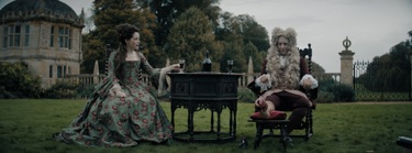 A still from the film 'Savage House'. A man and a woman dressed in grand 18th century clothing sit on dark wooden dining chairs either side of a dark wooden table with wine, outside on a country estate
