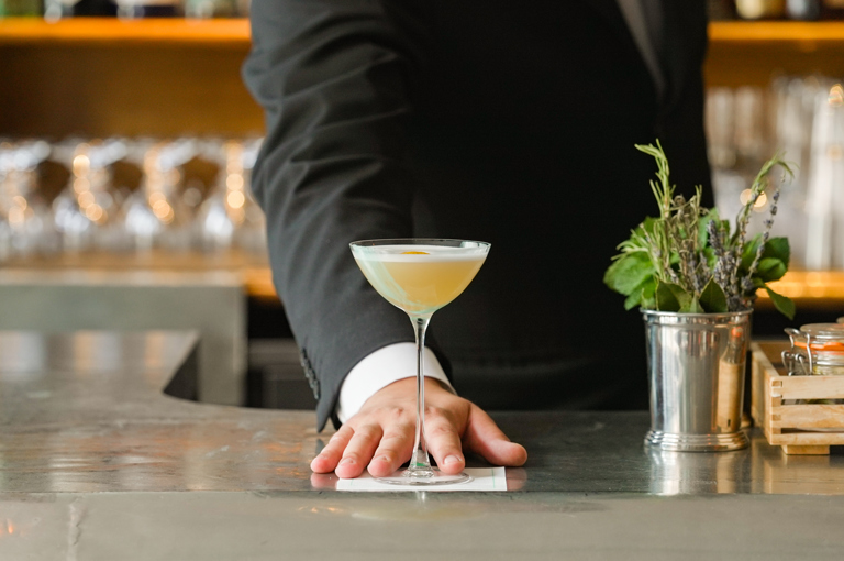 A t shot of the cocktail 'Grant's Palette' at Oscar Bar & Restaurant: the cocktail is being pushes forward by a bartender standing behind the bar. 