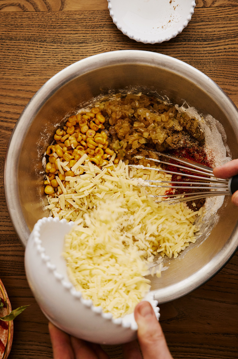 In this image you can see a large silver bowl filled with the ingredients to make the cornbread. on the left a hand is pouring in cheese from a white bowl and on the right the hand is whisking the ingredients together. 