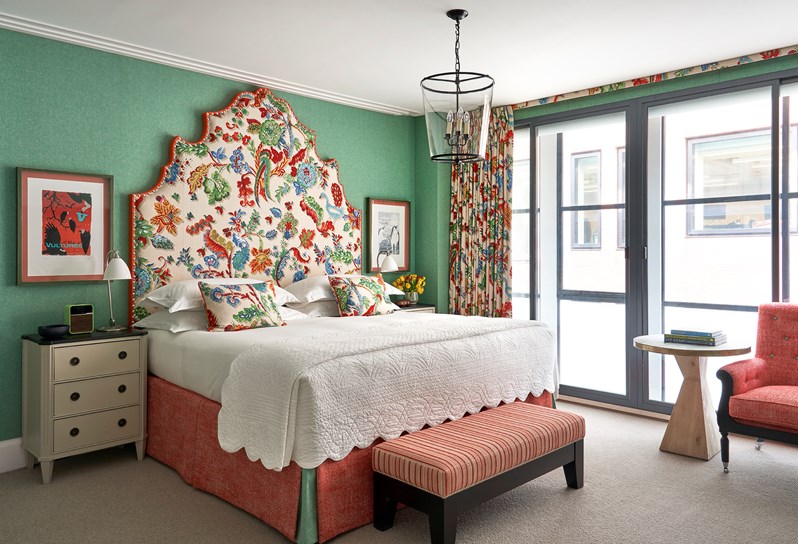 A bedroom with green wallpaper, featuring a king sized bed with a bold headboard in colors and a pattern inspired by nature and there is an ottoman at the end of the bed and a red armchair at the end of the room