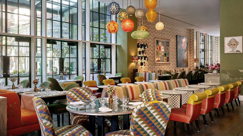 Diagonal shot of the Crosby Bar with a round table set up for 4 at the front and some bar tables with sofas and low chairs behind. View of a ceiling high curtain wall on the back left and of some of the restaurant tables on back right.
