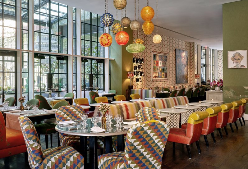 Diagonal shot of the Crosby Bar with a round table set up for 4 at the front and some bar tables with sofas and low chairs behind. View of a ceiling high curtain wall on the back left and of some of the restaurant tables on back right.
