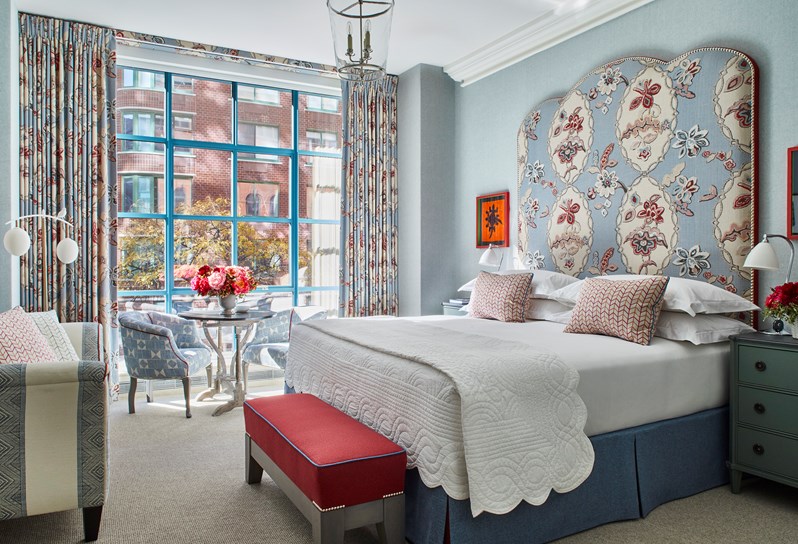 Bed with a blue and pink patterned headboard and matching curtains, a red ottoman, two blue patterned armchairs, a coffee table and a blue and white striped sofa
