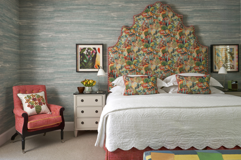 A front on image of the bedroom of The Deluxe Two Bedroom Suite, Room 5. A stylish bedroom featuring a vibrant patterned headboard and pillows, blue textured wallpaper, eclectic artwork, and an arm chair in the room.