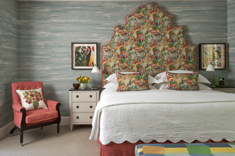 A front on image of the bedroom of The Deluxe Two Bedroom Suite, Room 5. A stylish bedroom featuring a vibrant patterned headboard and pillows, blue textured wallpaper, eclectic artwork, and an arm chair in the room.