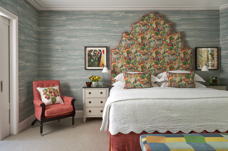A front on image of the bedroom of The Deluxe Two Bedroom Suite, Room 5. A stylish bedroom featuring a vibrant patterned headboard and pillows, blue textured wallpaper, eclectic artwork, and an arm chair in the room. 