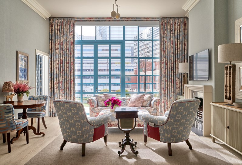 A living room area. Two blue and white patterned armchairs in front of footstall in blue 'Chainstitch' fabric which matches the sofa opposite and curtains on the floor to ceiling windows behind. On the left, two blue patterned chairs with a wooden table in the middle and a door open to the next room. On the right, a TV, fireplace, two lamps and a side cabinet.