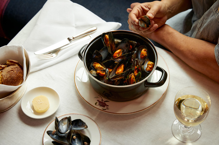 A hand picks a mussel from a large black pot of steamed mussels with a side of bread at Brumus