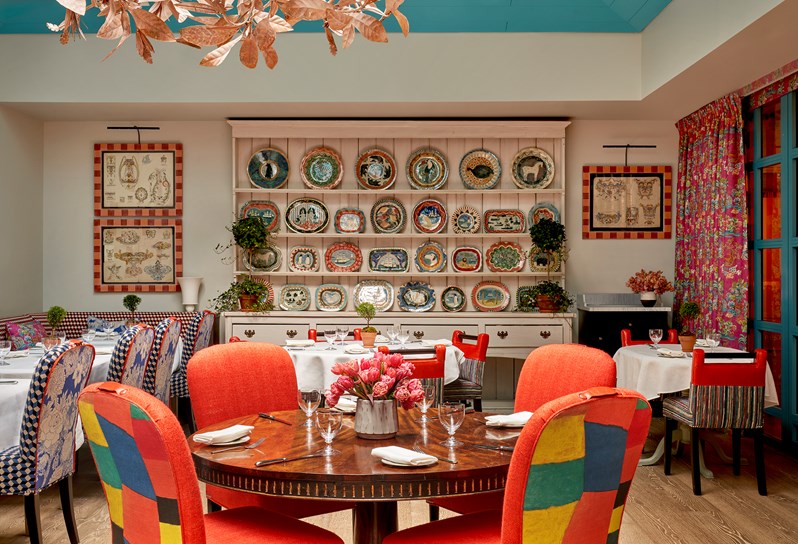 A round wooden table with four bright orange chairs in The Orangery at Warren Street Hotel. In the background, a large painted dresser is filled with ceramic plates