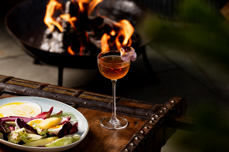 A cocktail and a dish of fire-cooked food sit on a wooden coffee table with a fire bowl in the background