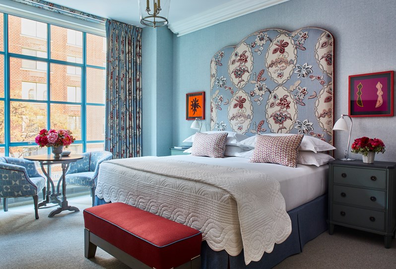 Bed with a blue and pink patterned headboard and matching curtains, a red ottoman and two blue patterned armchairs