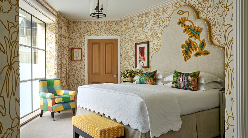 A bedroom with walls decorated in a textured fabric with a yellow leaf pattern and a large bed with a decorative headboard. A yellow footstool sits at the end of the bed and to the left is a small chair upholstered in a multicoloured fabric
