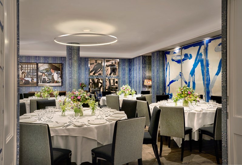 An image of multiple round dining table set up in the Indigo Room, there are white table cloths, floral bouquets in the centre and candles. each place around the table has a placemat, plate, cutlery, glassware and napkins ready for dining. private events room at The Soho Hotel.