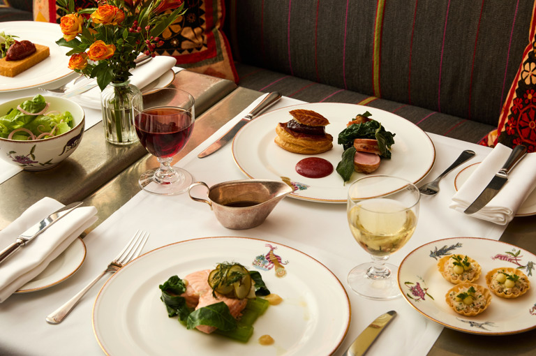 A close up image of a table at Refuel at Soho Hotel with white linen clothes is set up with multiple dishes from the Summer A La Carte with glasses of red and white wine on each place setting. 
