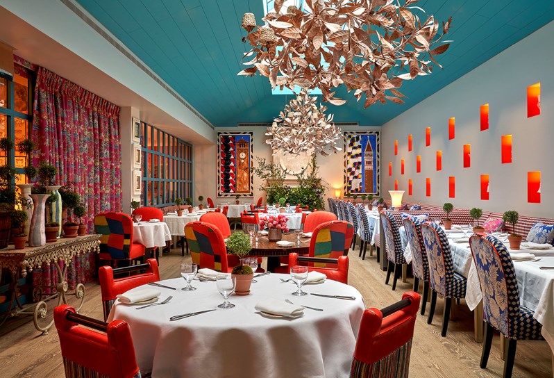 The Orangery at Warren Street Hotel with a vaulted blue ceiling and skylights. Banquette seating runs down the left-hand side of the room and several round tables are dotted the length of the room. Two statement chandeliers hang from the ceiling and at the end of the room is a fireplace filled with foliage