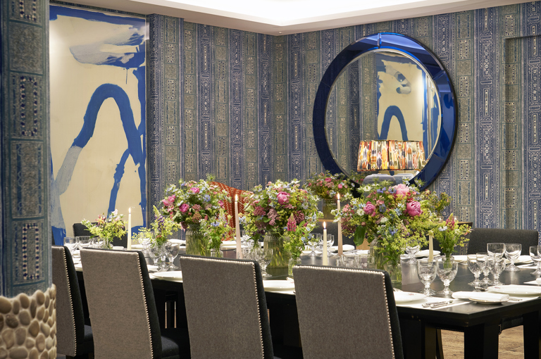 An image of the long dining table set up in the Indigo Room, there are floral bouquets in the centre and candles. each place around the table has a placemat, plate, cutlery, glassware and napkins ready for dining. private events room at The Soho Hotel.