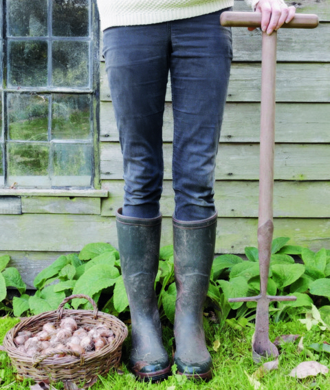An image to promote Harriet Thystlethwaytes new book 'A country style: living and creating with nature through the seasons'. Image shows Harriet's legs with a wicker basket off bulbs on the left and she is holding a spade.