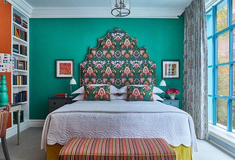 A bed with a statement turquoise headboard and cushions with a multi-coloured pattern and a striped ottoman, two square prints on the wall either side of the bed. A bookshelf and mirror on an orange wall with a desk, turquoise lamp and striped chair.