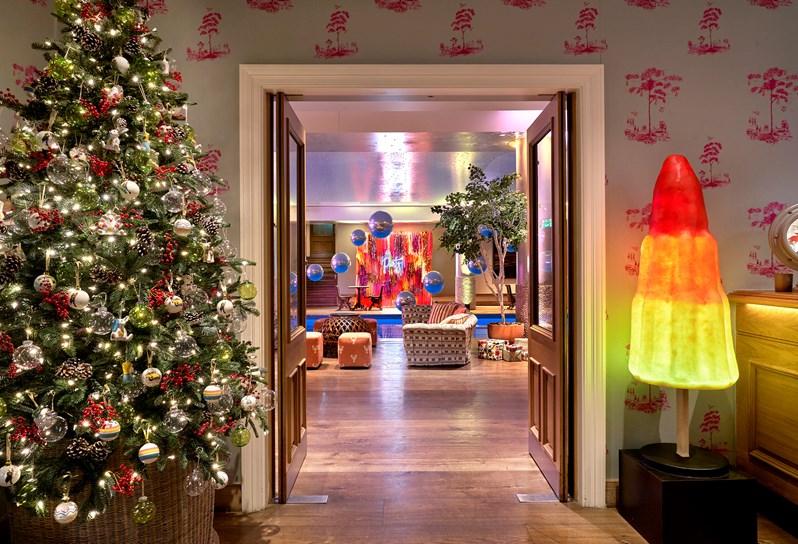 The entrance to the Pool area with a Christmas tree on the left hand side and a three coloured iceberg - red orange and yellow. The pool area can be seen with large metallic looking balls hanging from above alongside a sofa are next to it