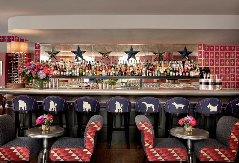 The bar at Brumus in Haymarket Hotel. Bar stools embroidered with dogs line the bar. In front there are two low tables with pink patterned seats either side.