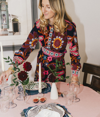 Artist and curator Domenica Marland dressed in a colourful patterned dress, creating a table scape. She is placing a candlestick on a dining table dressed with a pink tablecloth, plates, glasses, tablemats and small bud vases