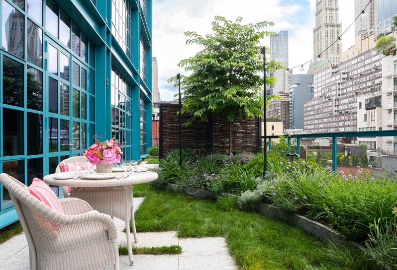 A terrace in the city with a white wicker table and chairs decorated with coral cushions, sitting on a patio. They are surrounded by grass and several plant pots filled with greenery line the edge of the balcony.