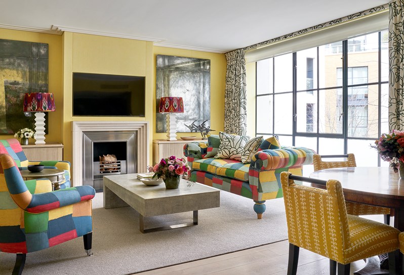 A living area of a suite. The room has yellow walls. There is a TV on the wall with a fireplace below, there are tables with lamps on either side. A sofa and two armchairs are upholstered in a multi-coloured 'Zig Zag' pattern. On the right, there is a dining table with yellow chairs.