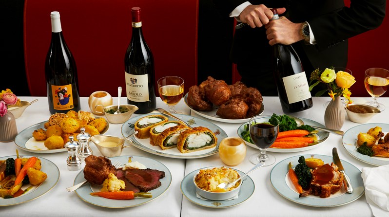 Table with plates of roast dinner with side dishes of cauliflower cheese, roast potatoes and vegetables. There are three magnums of wine, one is being opened by a man in a suit