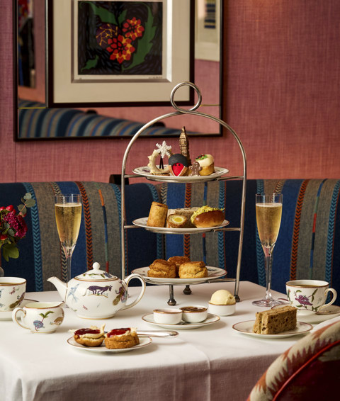 This is a portrait image of the Christmas London Calling festive afternoon tea offering at Brumus at Haymarket Hotel. This image shows a table in Brumus set with Mythical creatures crockery and the full afternoon seat set up including pots of tea, tea cups, glasses of champagne and a stand with sandwiches, scones with preserves and cakes all in a festive and classically British theme.