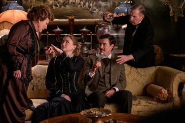 A still from the film 'Virginia Woolf's Night and Day': the image displays four people enjoying champagne in a living room. The younger couple are sat on the patterned sofa while the older pair are standing