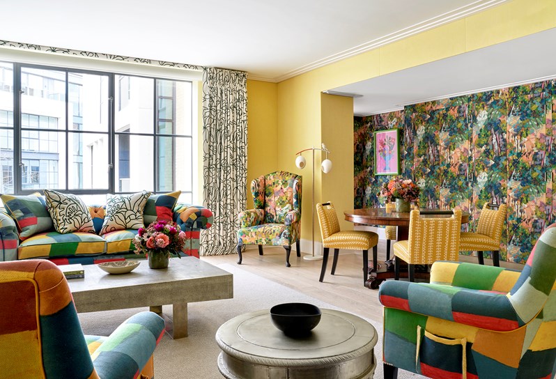 A living room space with a multicoloured sofa and armchairs and a rectangular coffee table. A round wooden table sits in in the right hand corner with yellow chairs