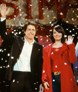 A still from 'Love Actually'. A young man and woman stand next to eachother on a stage, smiling and waving as confetti falls around them