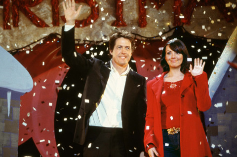 A still from 'Love Actually'. A young man and woman stand next to eachother on a stage, smiling and waving as confetti falls around them