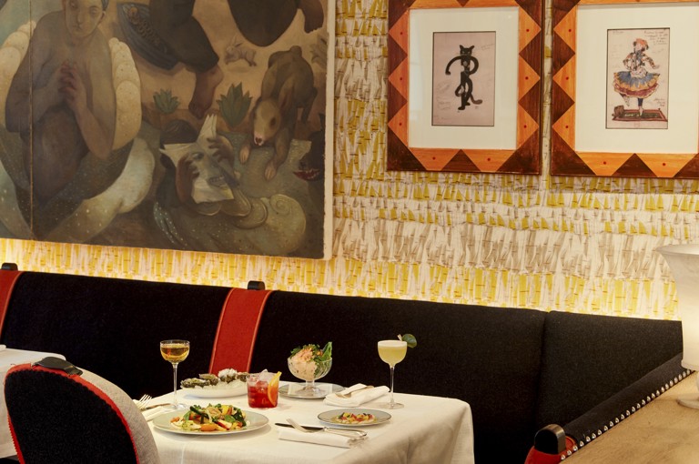An image to promote the Summer à la carte starters at Ham Yard Bar & Restaurant at Ham Yard Hotel. The image shows a white-linen table set with a selection of starters, including oysters, prawn cocktail, and vibrant salads. Three cocktails are placed alongside the dishes, set against a backdrop of patterned wallpaper and framed artwork