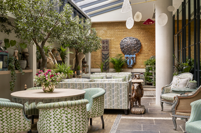 An image of the orangery at the Ham Yard Restaurant, Ham Yard Hotel. The image shows the round table in the foreground of the image with four tub chairs in Little Weed in green around it. Behind that table we see two seating areas both with two sofas facing each other. 