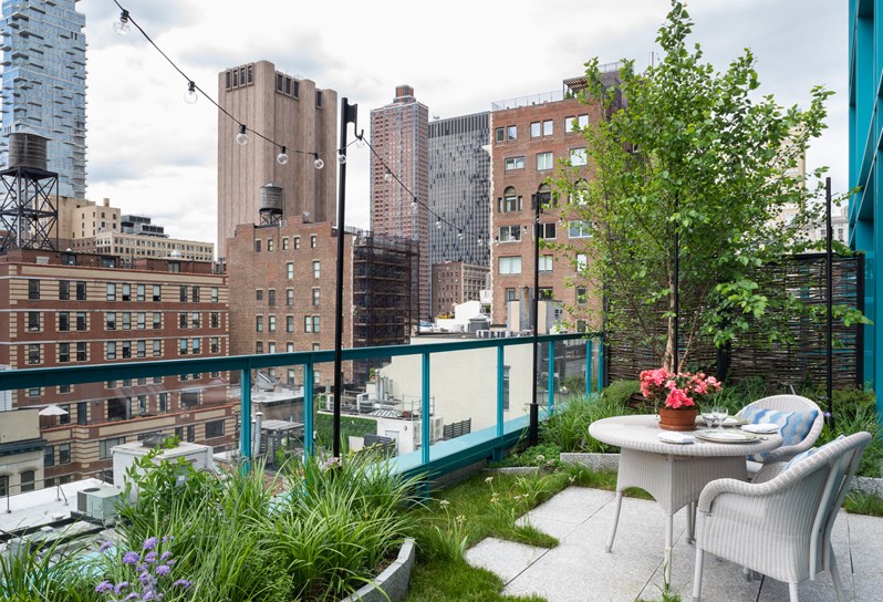 A wraparound landscaped terrace balcony. Plants and grass around the outskirts and a small patio in the middle with white woven outdoor dining chairs and table.